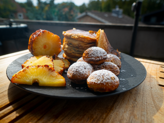 Auf einem schwarzen Teller liegt eine Auswahl an Speisen: karamellisierte Ananasscheiben mit Röstaromen, ein Stapel dicker Pancakes und eine Portion runder, mit Puderzucker bestreuter niederländischer Poffertjes (kleine Pfannkuchen). Der Teller steht draußen auf einem Holztisch, im Hintergrund sind Bäume und ein Hausdach zu sehen.