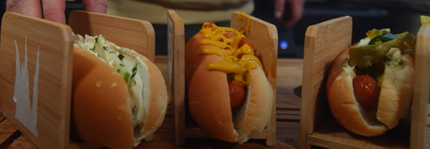 Drei Hot Dogs in Brötchen stehen nebeneinander auf einem Holzbrett, jeder in einem eigenen Holzhalter. Der linke Hot Dog ist mit einem cremigen Belag und Kräutern gefüllt, der mittlere mit Senf überzogen, und der rechte enthält eingelegte Gurken und Jalapeños.